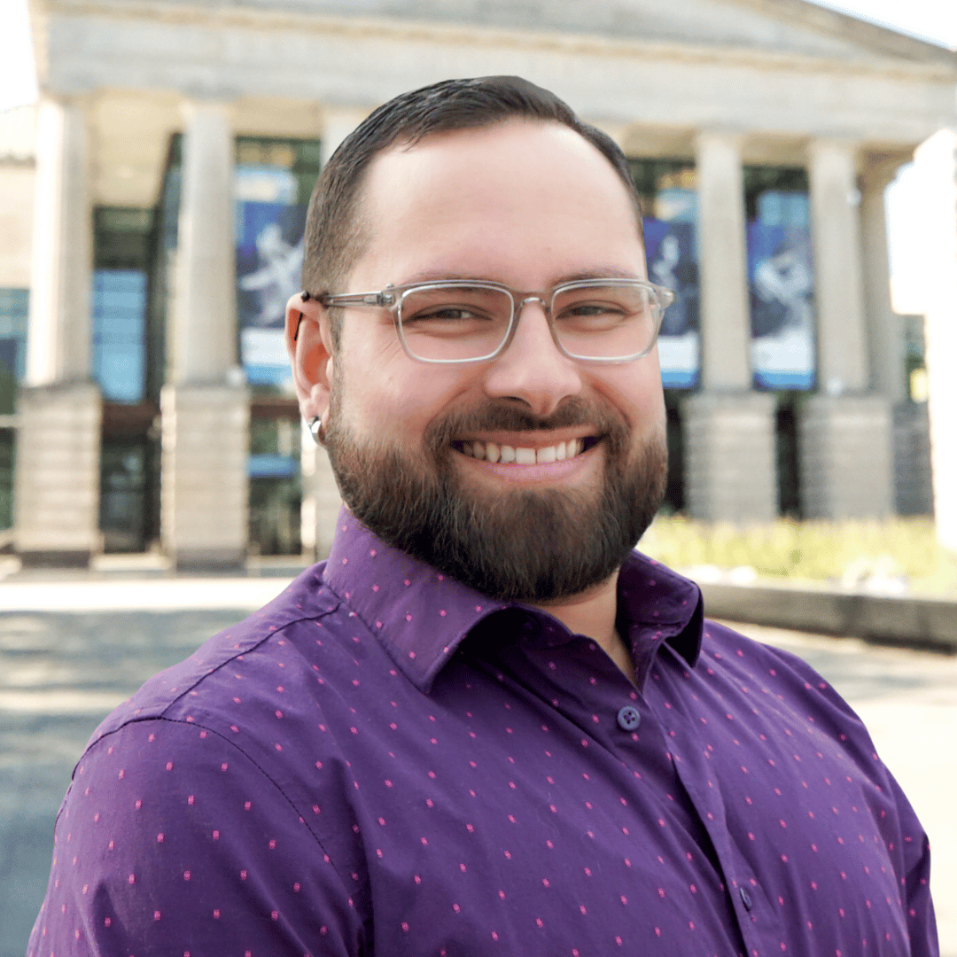 Headshot of Andrew Armas in front of the Martin Marietta Center for the Performing Arts wearing a purple button down and glasses. 
