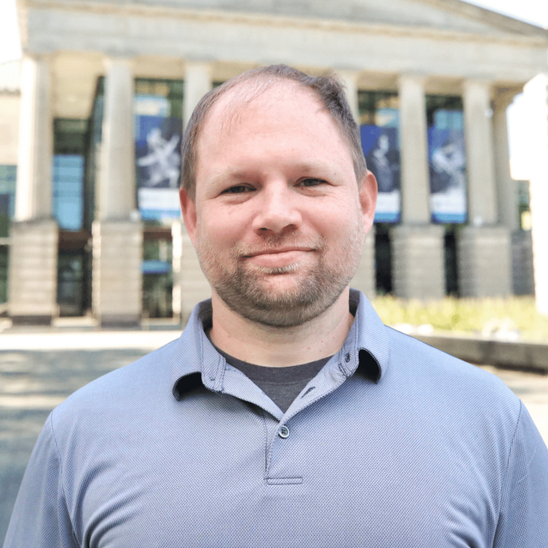 Headshot of Brian Moore in front of the Martin Marietta Center for the Performing Arts wearing a grey button up shirt.