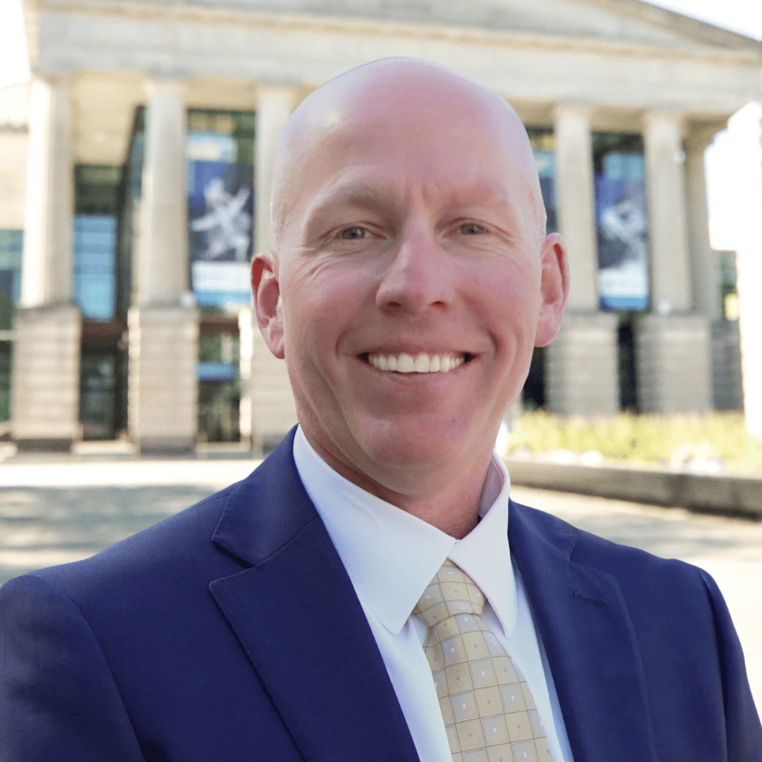 Headshot of Chris Bullock outside the Martin Marietta Center for the Performing Arts wearing a navy blue suit with a white button down and tie.