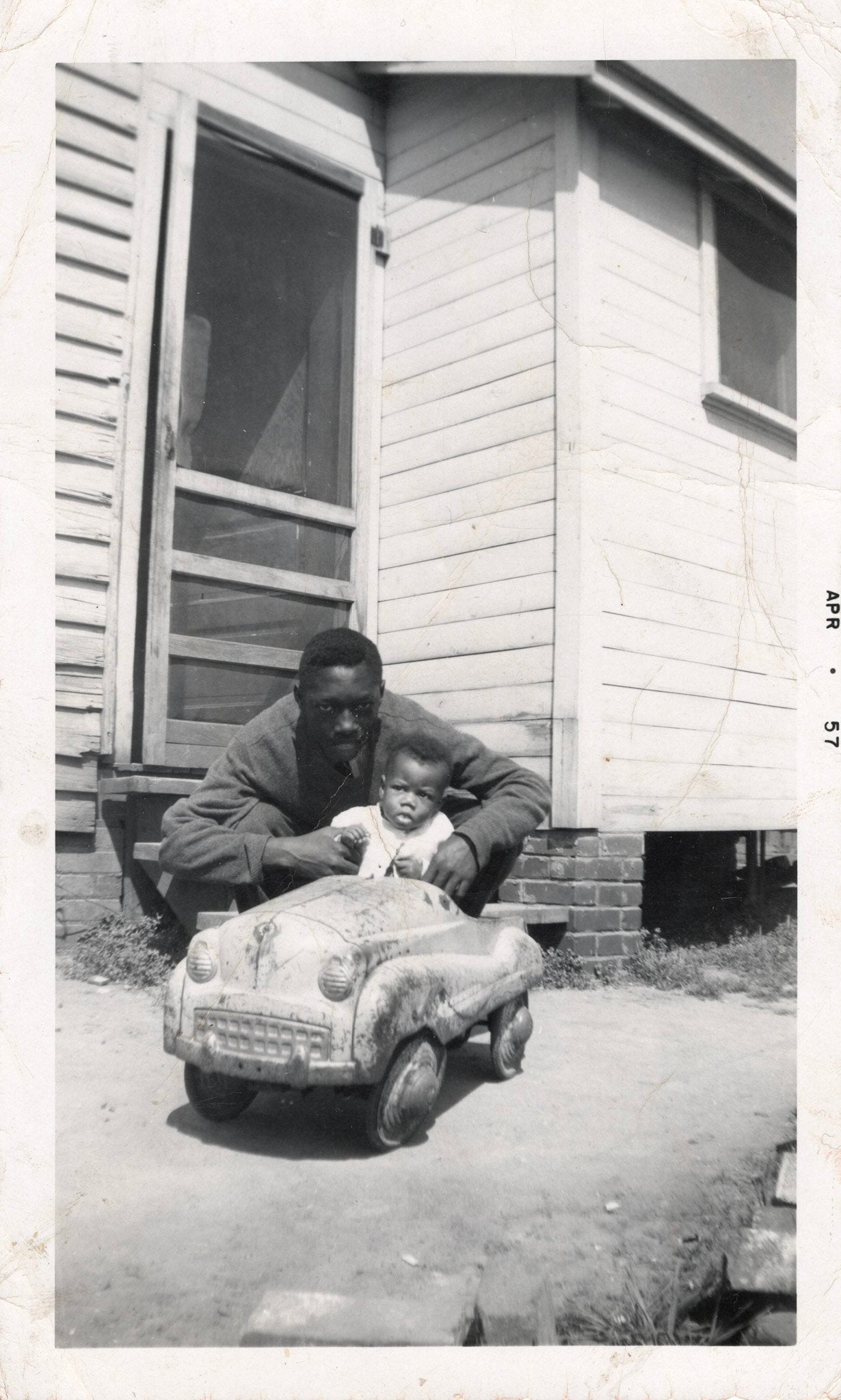 A photo of a young man he is squatting down behind a young child who is sitting in a toy car. a door to a house can be seen in the background.