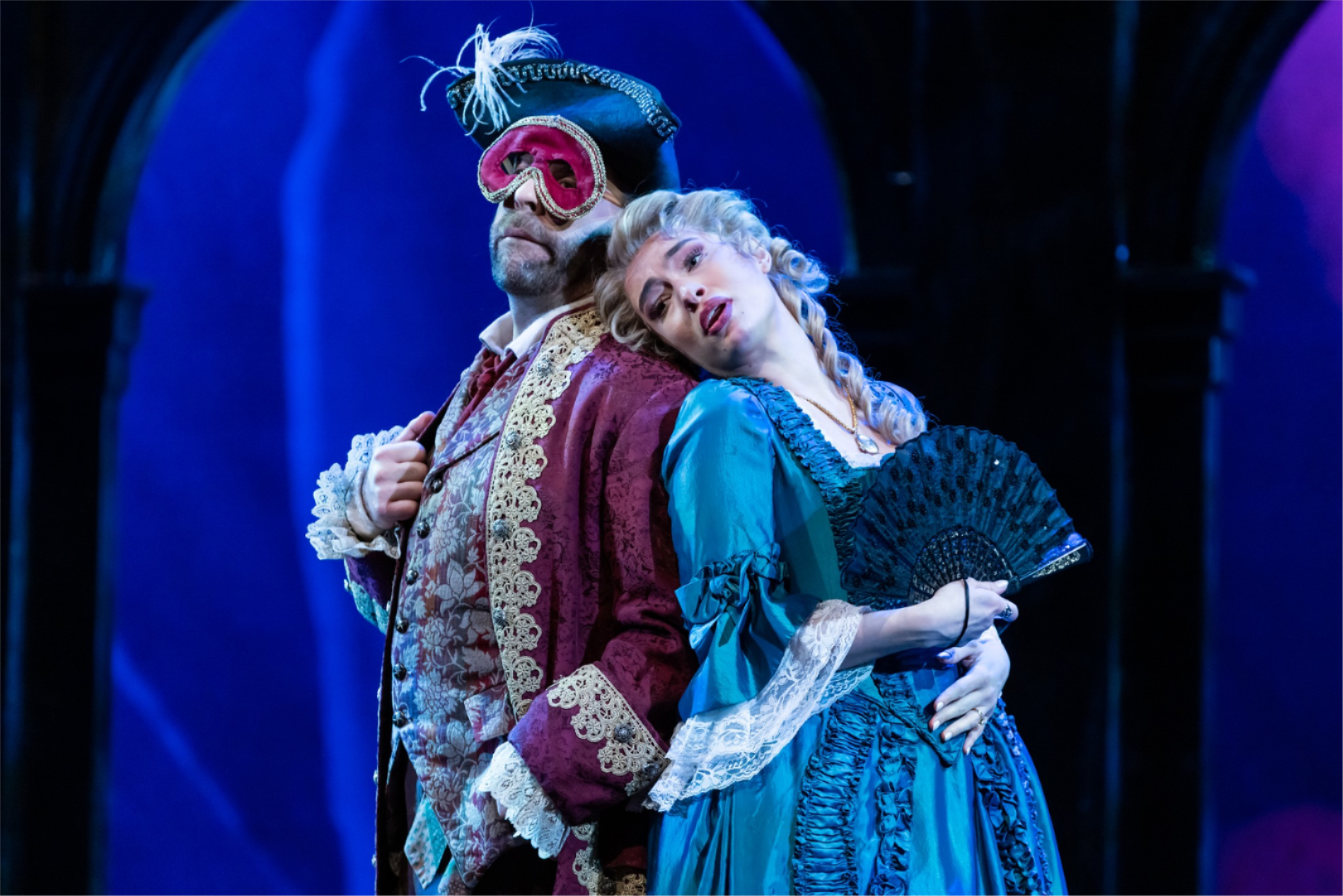 Two NC Opera performers shown in 1700s attire in the production of Don Giovanni the man is wearing a mask and hat and the woman is holding a fan