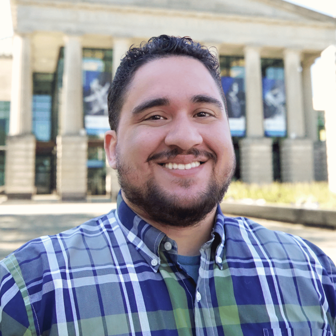 Headshot of EG Garcia in front of the Martin Marietta Center for the Performing Arts wearing a green and blue plaid button down shirt. 