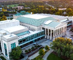 Exterior photo of Raleigh Memorial Auditorium with a green roof, classical columns, and glass windows, surrounded by trees and walkways.