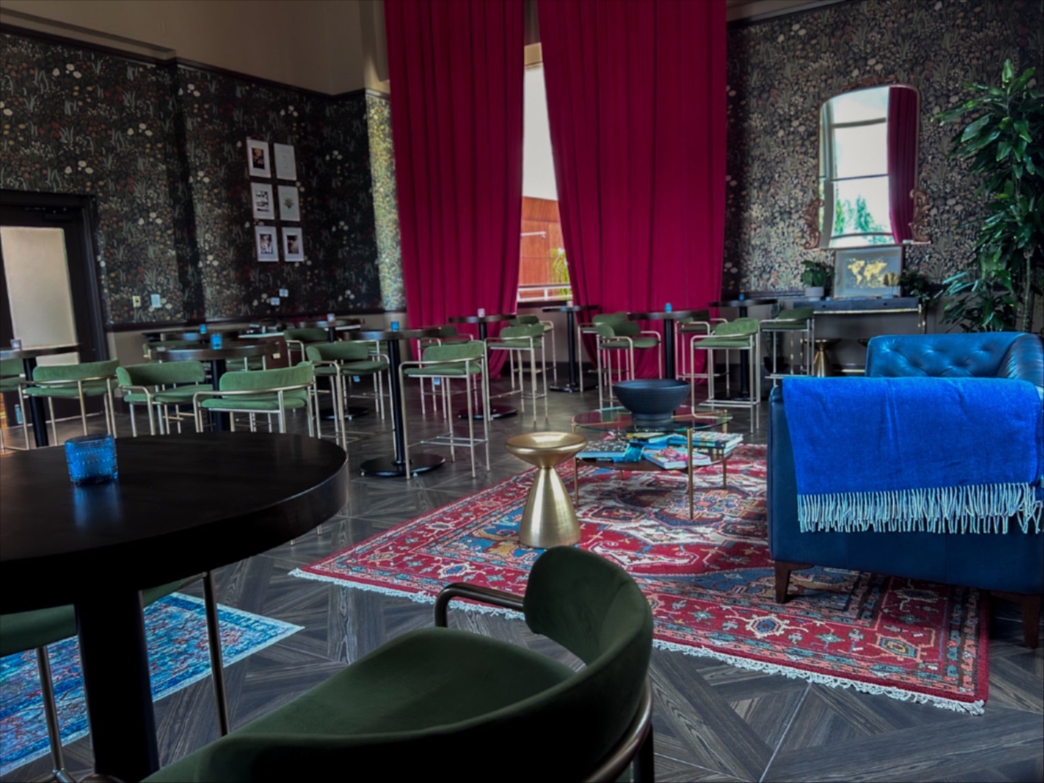 Stylish lounge with patterned wallpaper, red curtains, green chairs, a blue armchair, and colorful rugs creating a cozy, eclectic atmosphere.