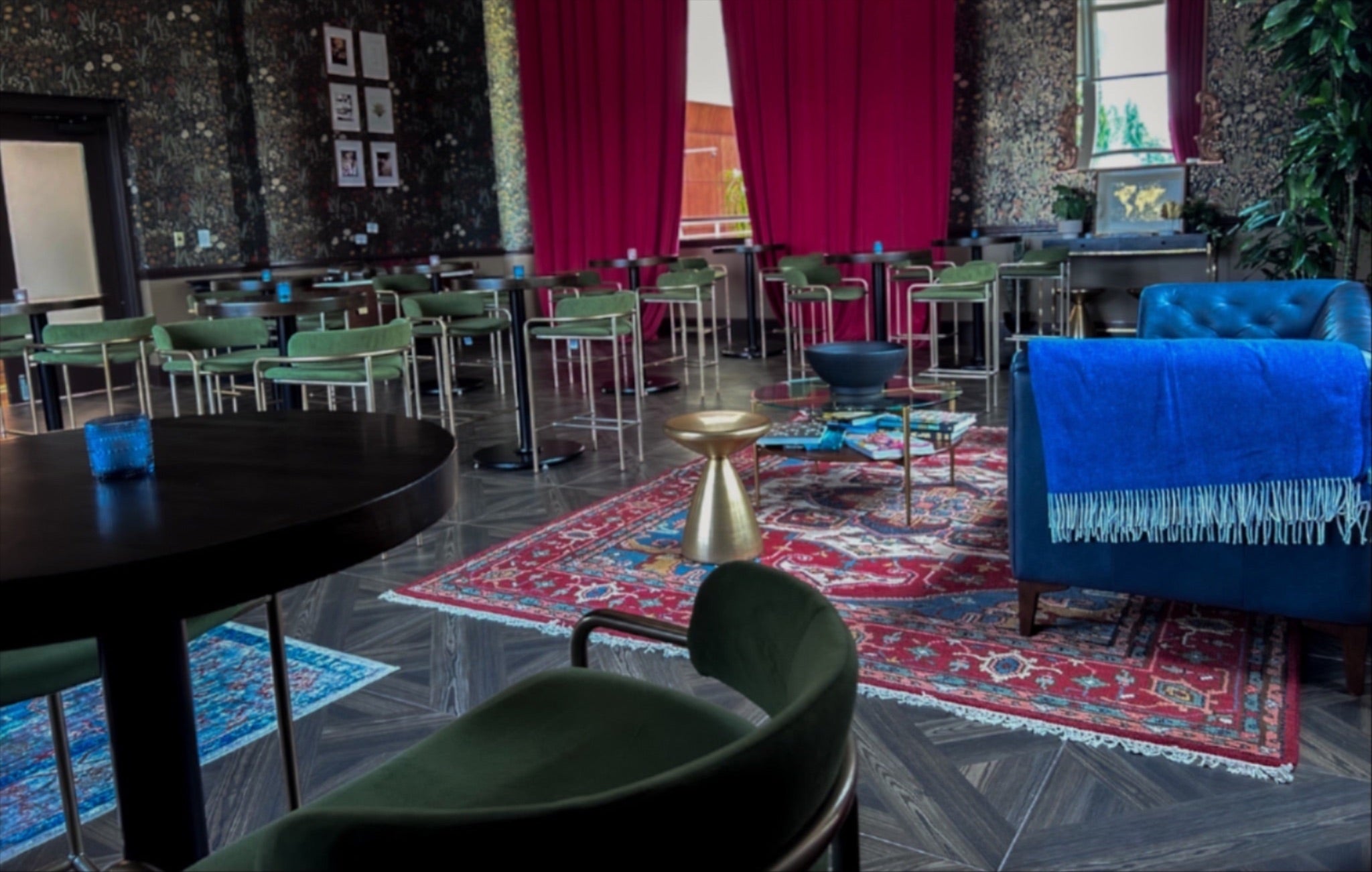 Stylish lounge with patterned wallpaper, red curtains, green chairs, a blue armchair, and colorful rugs creating a cozy, eclectic atmosphere.