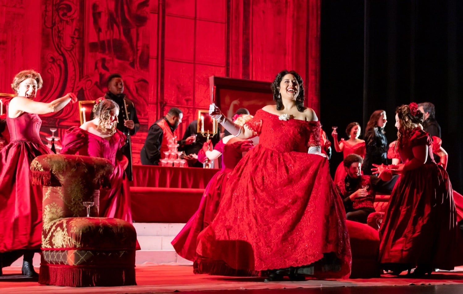 An opera singer is shown from the production La Traviata she has dark hair and is wearing a bright red long dress