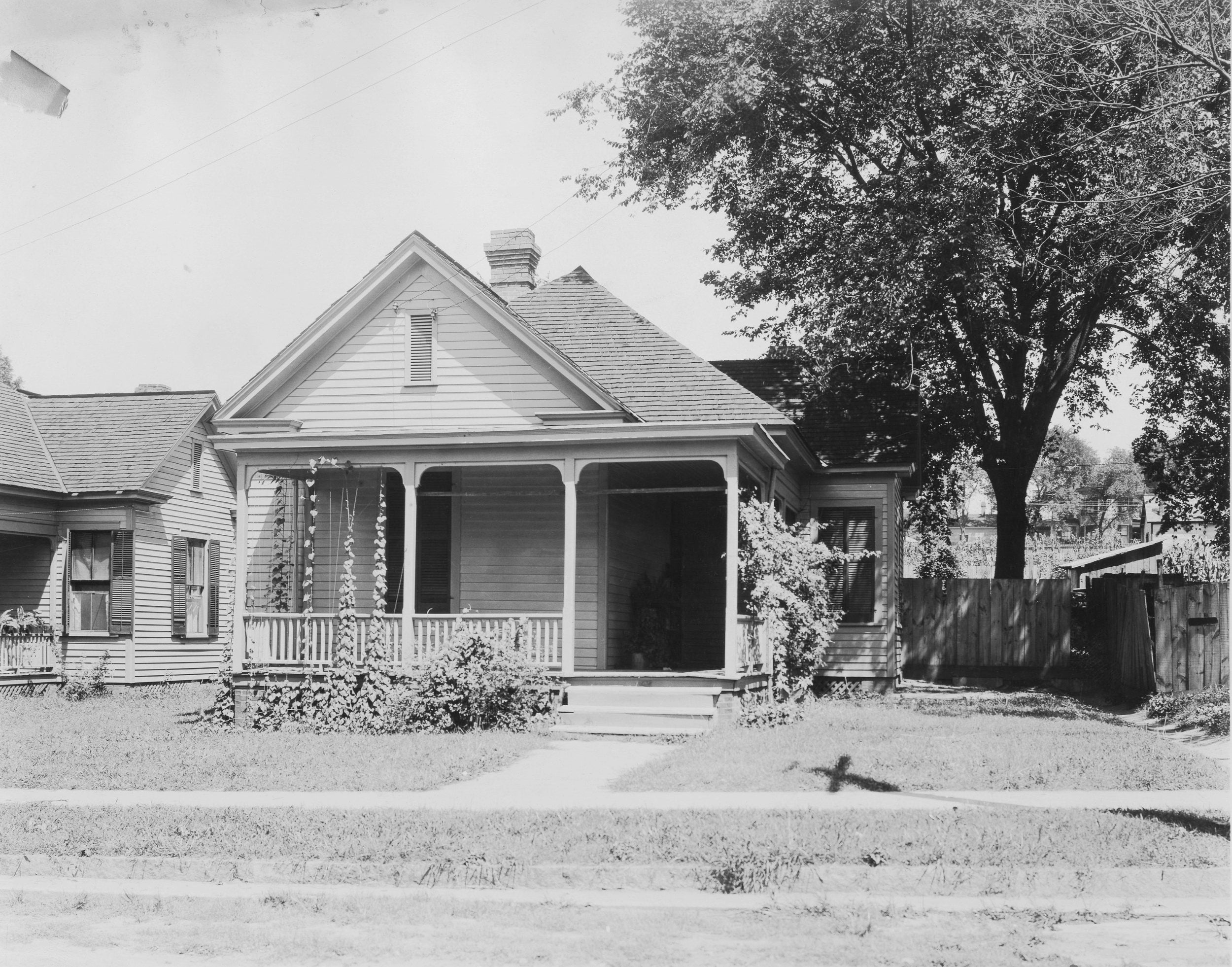 a black and white photo of a home is shown. there is a tree and fence to the right of the home and another home on the left side.