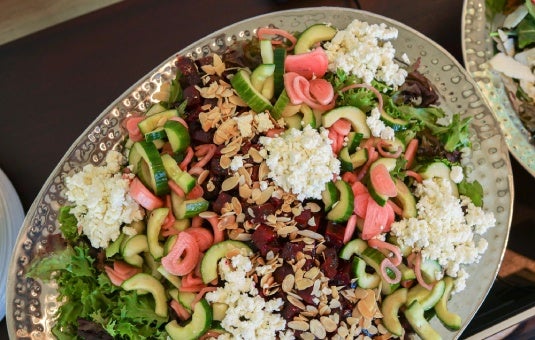 Colorful salad with mixed greens, sliced cucumbers, pickled vegetables, crumbled cheese, and slivered almonds on a large silver platter.