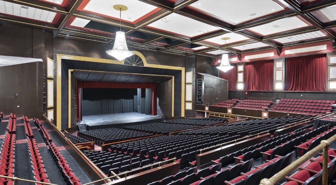 Interior view of Raleigh Memorial Auditorium with red and black seating, chandeliers, and a stage framed by gold and black trim.