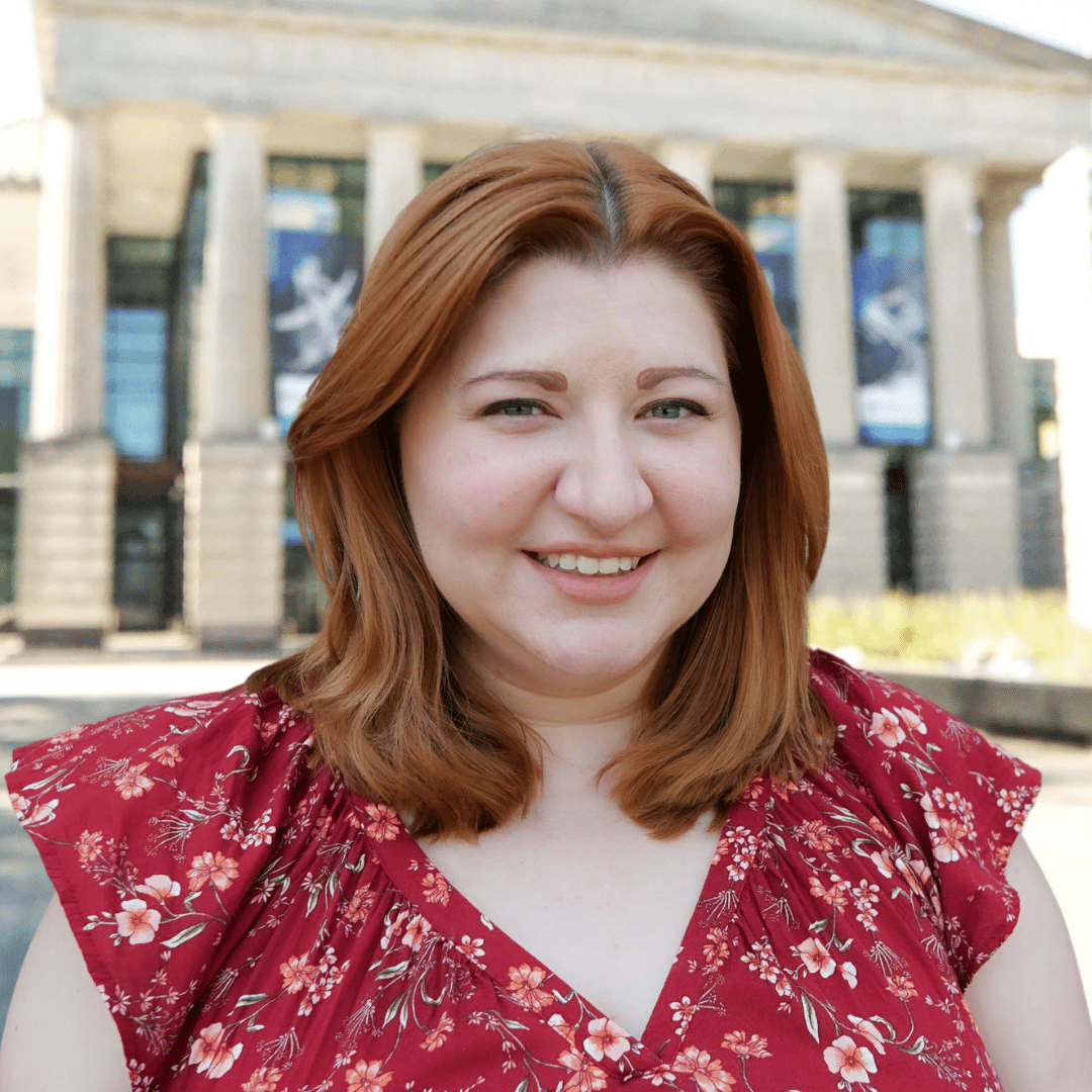 Headshot of Nicole Ackman in front of the Martin Marietta Center for the Performing Arts wearing a red floral blouse. 