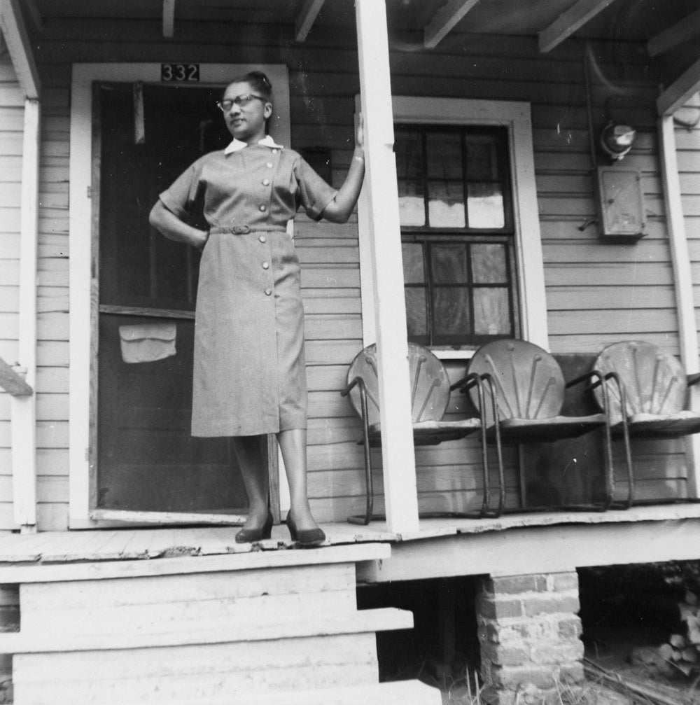 a black and white photo of a lady standing on a front porch her left arm is raised leaning against the porch post she is wearing a dress and classes looking to her right.