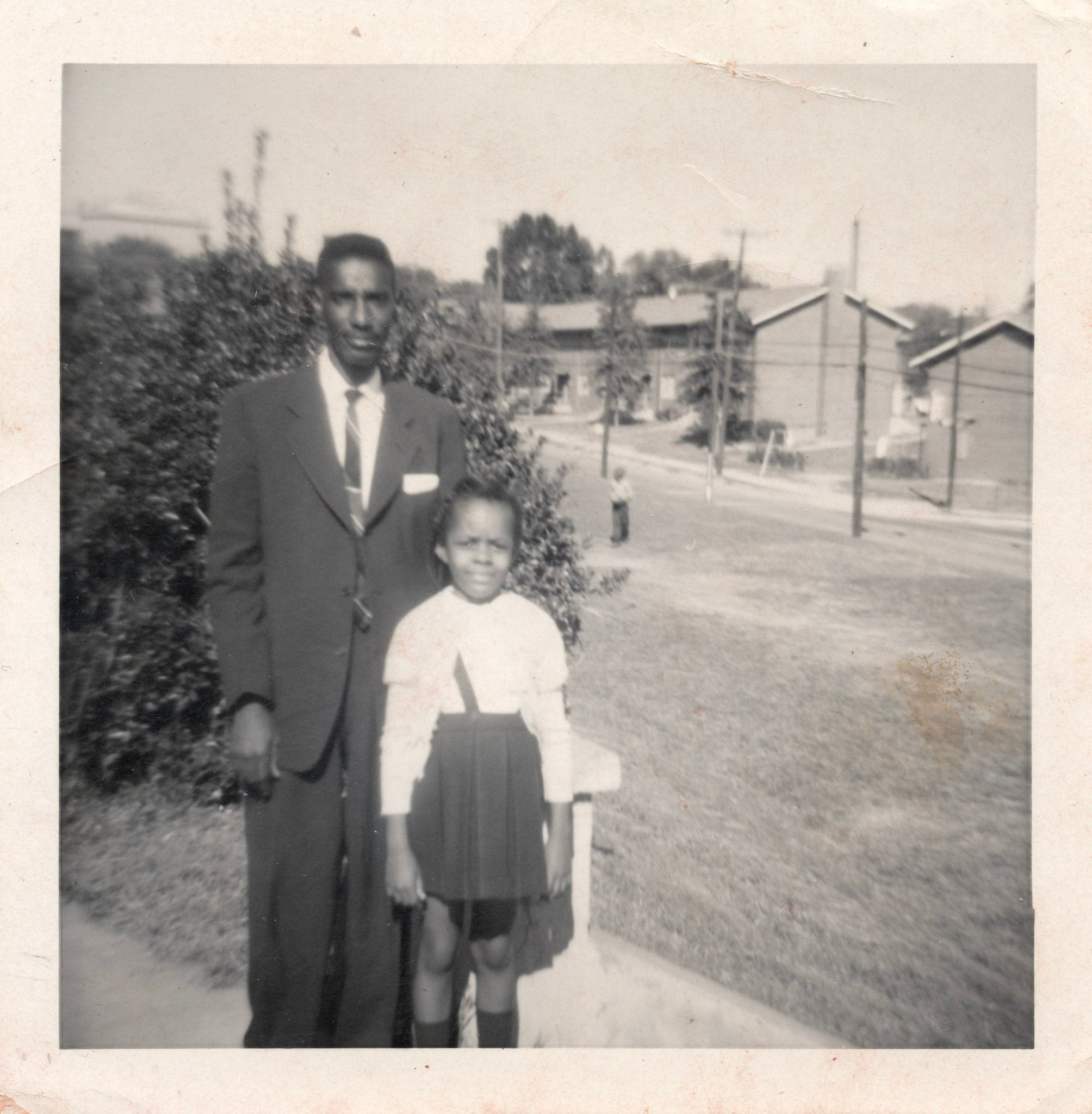 a black and white photo of a man and young girl. he is wearing a suite and tie and the girl is wearing a skirt and blouse. they are standing in front of foliage, you can see a field and houses behind them.
