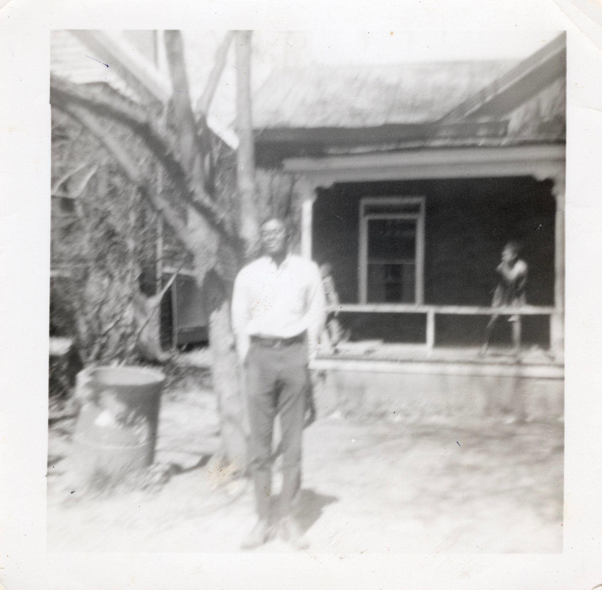 a black and white photo of a young man he is standing in front of a house on the porch is a young child leaning on the railing.