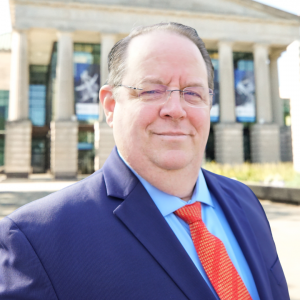 Headshot of Rob Leavell in front of the Martin Marietta Center for the Performing Arts wearing a blue suit with a red tie. 