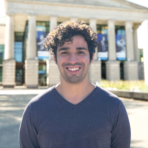 Headshot of Sean Delgado-Cruz in front of the Martin Marietta Center for the Performing Arts wearing a dark grey sweater. 
