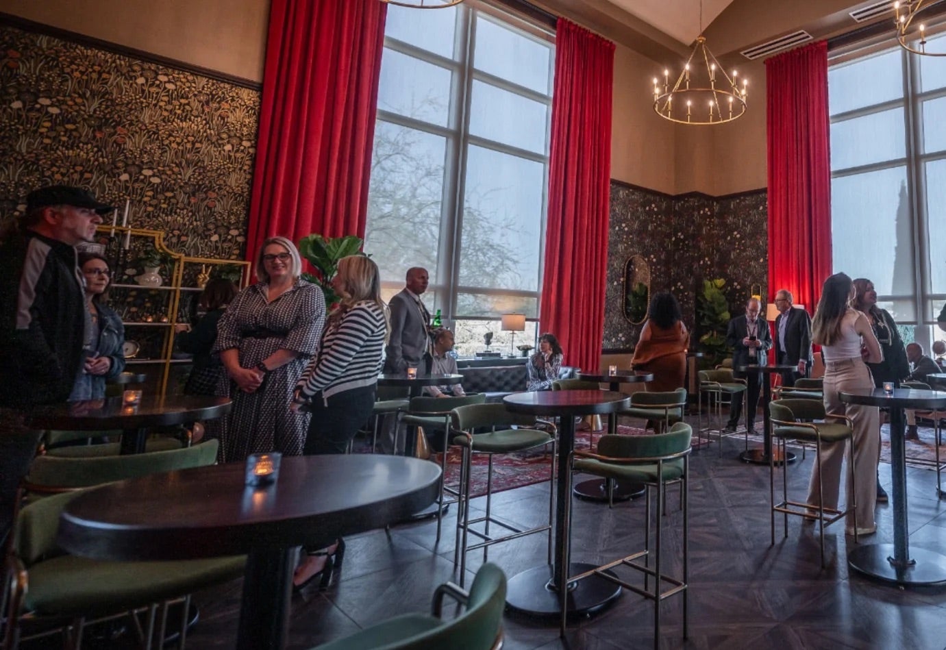 a photo of a hospitality lounge. there are bright red curtains and large windows shown in the background. in the center of the room are people gathered around cocktail tables.