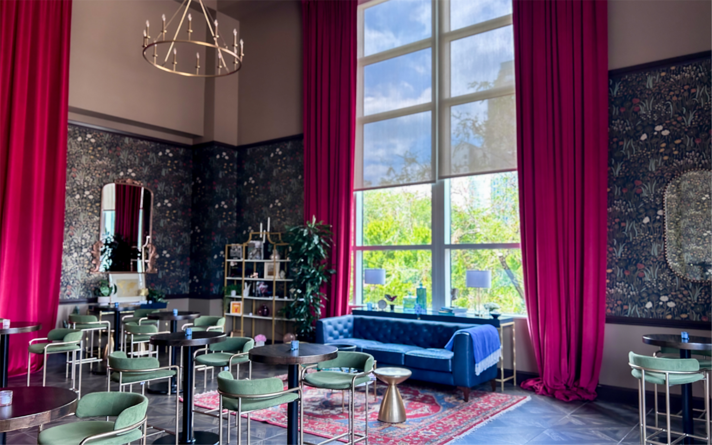 A photo of a lounge space a large window is shown on the right side with red curtains there is a blue couch under the window and high top tables to the left throughout the room