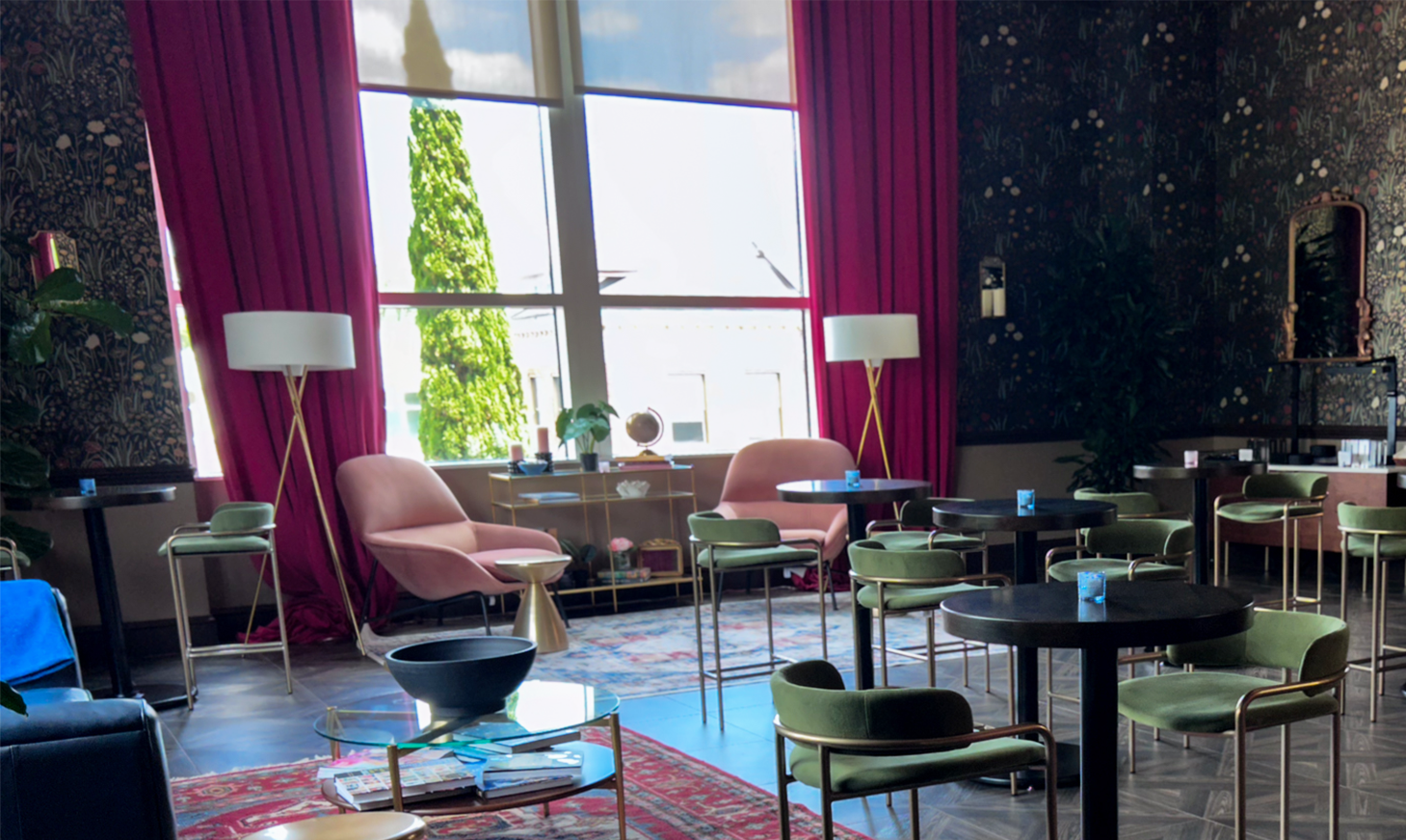 A photo of a lounge space there is a window with red curtains on the left side and high top tables shown throughout the room