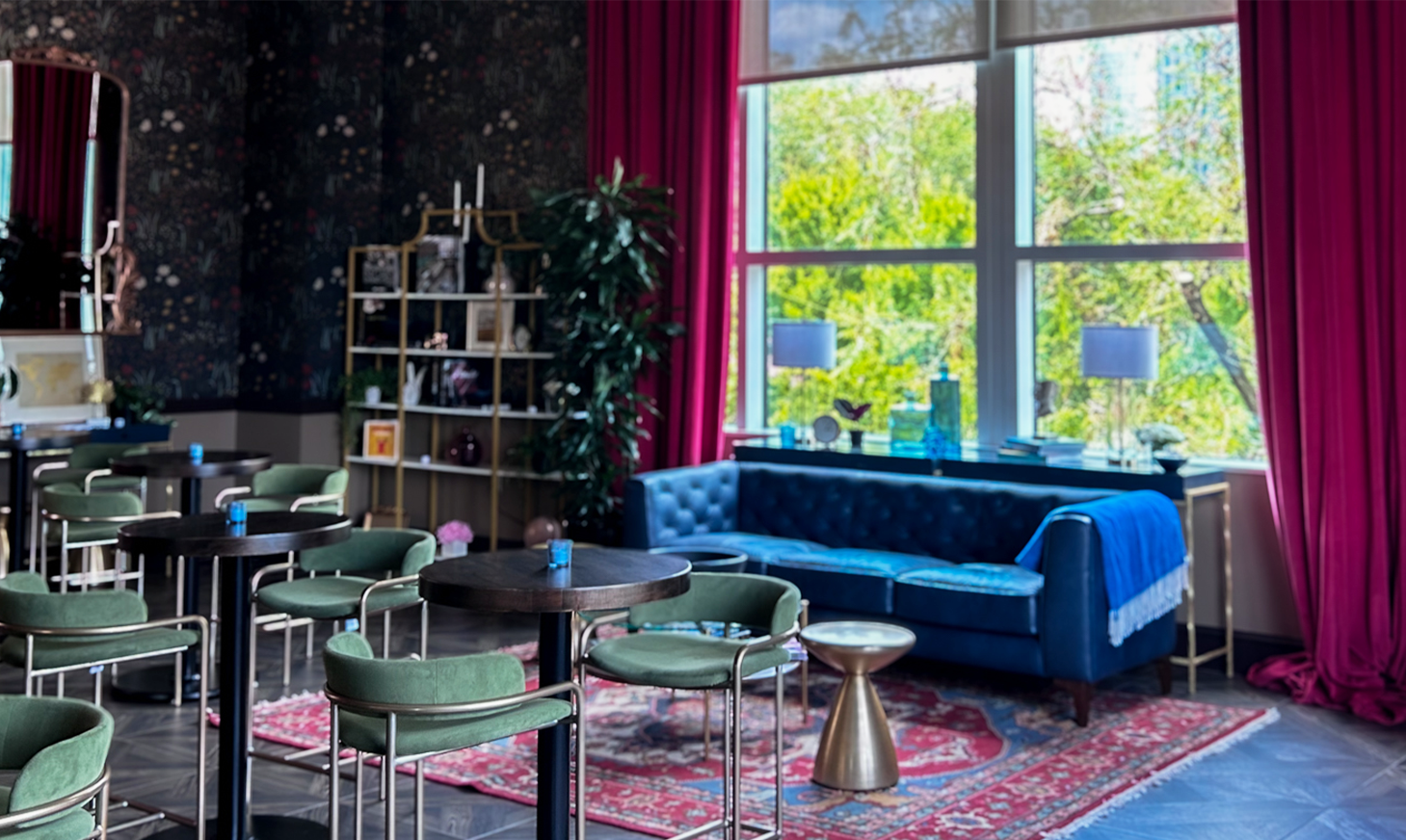 A photo of a lounge space there is a window with red curtains on the right side with a blue couch underneath the window and high top tables shown throughout the room