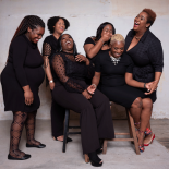 A group of six people, two sitting and five standing. They are dressed in all black, and are laughing.