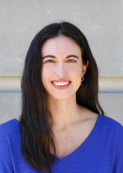 An image of Melanie Margarum, she is wearing a blue shirt and standing in front of a grey brick wall