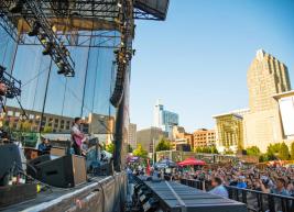 The left side of the photo shows a band on stage performing at the red hat amphitheater and the right side shows the raleigh skyline buildings and the audience