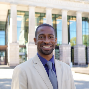a photo of Clifton simmons he has on purple button down shirt and purple tie with a tan sport coat