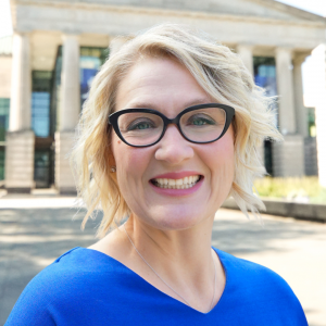 a photo of Michelle Bradley she has short blonde hair and glasses and is wearing a bright blue blouse