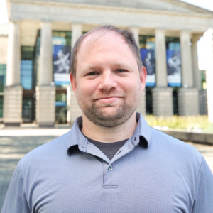 A photo of Brian Moore wearing a grey polo standing in front of Raleigh Memorial Auditorium .