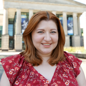 A photo of Nicole Ackman wearing a red blouse standing in front of Raleigh Memorial Auditorium .